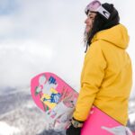 Jonge vrouw in gele jas met roze snowboard geniet van haar dure skipas in Italië