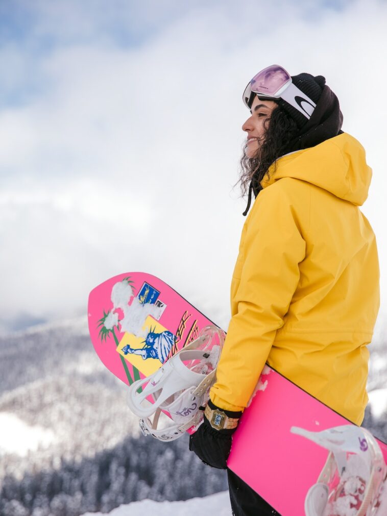 Jonge vrouw in gele jas met roze snowboard geniet van haar dure skipas in Italië