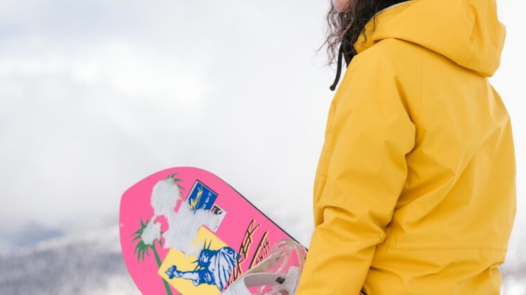 Jonge vrouw in gele jas met roze snowboard geniet van haar dure skipas in Italië